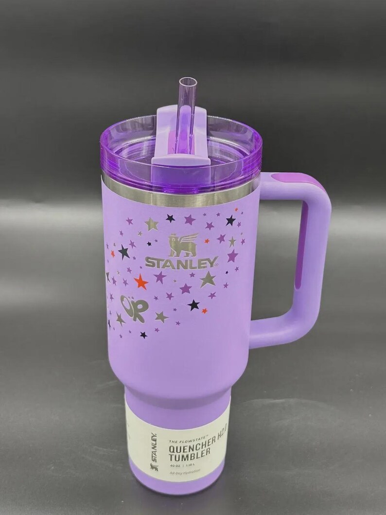 May include: A purple Stanley Quencher H2.0 Flowstate tumbler with a handle and a straw. The tumbler features a star pattern and the Stanley logo. The text "The Flowstate Quencher H2.0 Tumbler" is printed on the bottom.