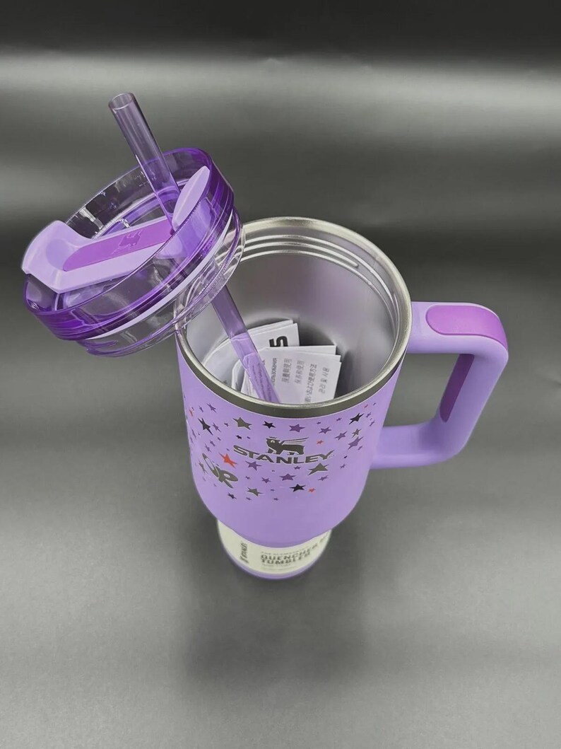 May include: A purple Stanley insulated mug with a handle and a clear lid with a straw. The mug has a star pattern and the Stanley logo. The lid is partially open, revealing the interior of the mug.