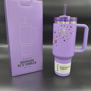 May include: A purple Stanley Quencher H2.0 tumbler with a handle and straw, next to its matching box. The tumbler is decorated with stars and a butterfly. The box and tumbler have the text "QUENCHER H2.0 TUMBLER" and "40 OZ | 1.18L".