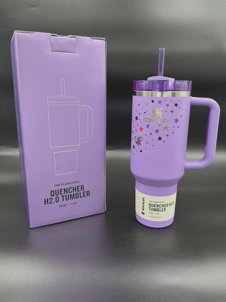 May include: A purple Stanley Quencher H2.0 tumbler with a handle and straw, next to its matching box. The tumbler is decorated with stars and a butterfly. The box and tumbler have the text "QUENCHER H2.0 TUMBLER" and "40 OZ | 1.18L".