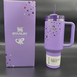 May include: A purple Stanley Quencher H2.0 tumbler with a handle and straw, next to a matching box. The tumbler and box are decorated with star graphics. The tumbler has the Stanley logo and the text "QUENCHER H2.0 TUMBLER".