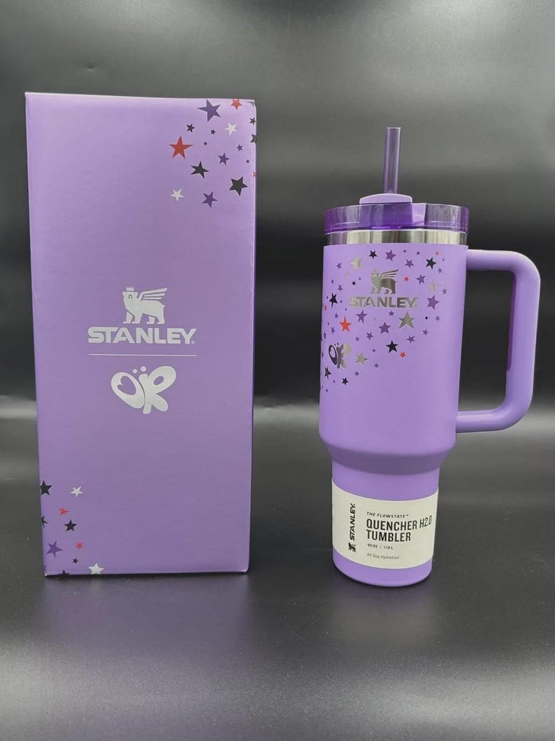 May include: A purple Stanley Quencher H2.0 tumbler with a handle and straw, next to a matching box. The tumbler and box are decorated with star graphics. The tumbler has the Stanley logo and the text "QUENCHER H2.0 TUMBLER".