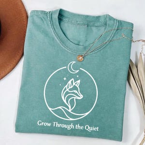 May include: A teal-colored t-shirt with a white graphic of a fox inside a circle with a crescent moon and stars. The text "Grow Through the Quiet" is below the graphic. A silver necklace with a round pendant is on the shirt.