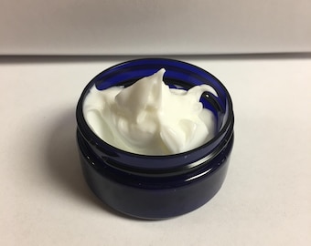 May include: A small, dark blue jar filled with a white, creamy substance. The jar is open, revealing the product inside. The cream has a thick, whipped texture, and the jar sits on a white surface.