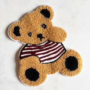 Puede incluir: Un parche de oso de peluche color canela con una superficie texturizada y difusa. El oso presenta detalles circulares negros en las orejas y los pies, una nariz negra bordada y una camisa blanca con rayas horizontales de color granate.
