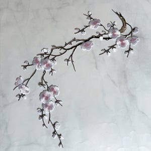 May include: An embroidered patch featuring a curved brown branch with multiple white cherry blossom flowers. Each flower has a soft pink center and delicate white petals. The branch extends across the frame with several smaller buds and twigs.