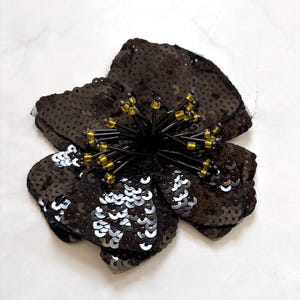 May include: A flower-shaped applique covered in dark brown and silver sequins. The center features long, thin black stems tipped with small, translucent yellow beads, creating a textured, layered appearance with a reflective, metallic finish.