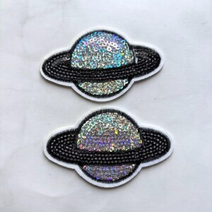 May include: Two embroidered patches in the shape of planets. Each patch features a shimmering, sequined planet body in shades of silver, blue, and purple, with a black sequined ring and a white border. The patches are designed for decorative use.