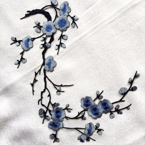 Puede incluir: Un parche bordado que presenta una rama negra oscura con flores y capullos en azul claro y azul oscuro. El diseño está dispuesto en una forma curva y fluida, mostrando un trabajo detallado de hilo en los pétalos y tallos sobre tela blanca.