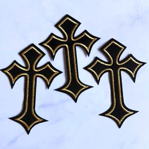 May include: Three embroidered patches in the shape of a cross. Each patch features a black center with a gold-colored stitched border. The crosses have pointed ends and a distinct, sharp-edged design, arranged in a cluster on a light surface.