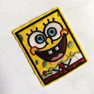 Op de afbeelding: Een rechthoekige geborduurde patch met een geel personage met grote witte ogen, blauwe irissen en een brede bruine mond met twee tanden. Het ontwerp bevat groene vlekken, een rode tong en een rood-witte rand op een gele achtergrond.