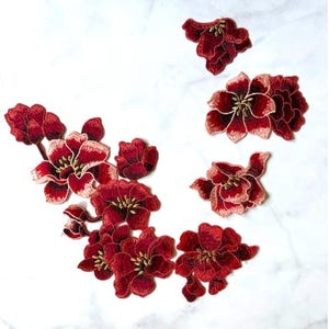 May include: A collection of embroidered flower patches in various shades of deep red, burgundy, and light pink. Each patch features intricate stitching with gold-coloured thread accents in the centre, depicting blooming flowers and buds.
