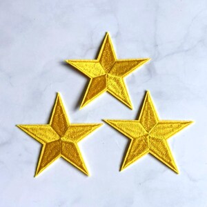Puede incluir: Tres parches idénticos en forma de estrella de cinco puntas con un acabado de hilo bordado en color amarillo vibrante. Cada estrella tiene un detalle de costura central distintivo que crea un sutil efecto tridimensional.