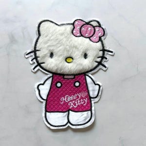 Puede incluir: Parche bordado con un gato blanco con lazo y vestido rosa. El parche tiene la inscripción "Heeey Kitty" en letras blancas. El gato tiene pelaje esponjoso y el diseño está delineado en negro.