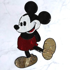 May include: A sequined iron-on patch depicting a cartoon mouse character. The design features a black head and ears, a white face, red shorts, and gold shoes. The sequins are arranged to create a shimmering, textured surface on the character's body.