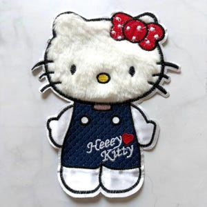 Puede incluir: Parche bordado con un gato blanco con un lazo rojo y un vestido azul marino. El parche tiene las palabras "Heeey Kitty" y un corazón rojo. El gato tiene pelaje esponjoso y el parche tiene un borde blanco.