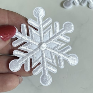 Könnte beinhalten: Ein weißer, bestickter Schneeflocken-Aufnäher mit einem sechsstrahligen Sterndesign und kreisförmigen Enden an jedem Arm. Die Stickerei besteht aus dichtem weißem Garn, das eine strukturierte, erhabene Oberfläche in Form einer Schneeflocke bildet.