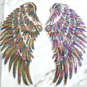 Könnte beinhalten: Zwei schillernde Engelsflügel-Patches mit Pailletten. Die Flügel bestehen aus kleinen, farbenfrohen Pailletten in Rosa-, Grün-, Blau- und Goldtönen. Die Flügel sind auf einer weißen Rückseite befestigt und zum Aufnähen auf Kleidung oder Accessoires bestimmt.