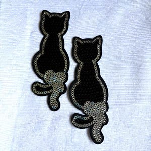 Puede incluir: Dos parches con forma de gato negro con detalles de lentejuelas. Cada gato está representado en posición sentada con un contorno de lentejuelas plateadas y un ovillo de lana de lentejuelas plateadas en la base de la cola. Superficie oscura y brillante.