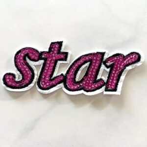 May include: A decorative iron-on patch featuring the word star in cursive script. The letters are filled with bright pink sequins, outlined in thick black embroidery thread, and bordered by a white fabric edge that follows the letter shapes.