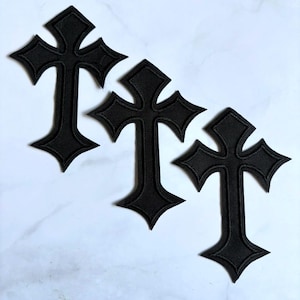 Puede incluir: Tres parches bordados en forma de cruz de color negro con bordes puntiagudos y un acabado de tela texturizada. Cada parche presenta una forma de cruz distintiva con brazos ensanchados y una punta inferior afilada, dispuestos en diagonal.
