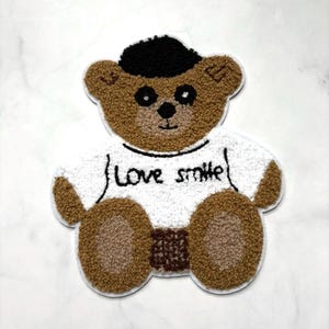 Peut inclure: Écusson en forme d'ours en peluche marron avec un chapeau noir et une chemise blanche. La chemise porte les mots "Love smile" en noir. L'ours a un visage et des pattes brun clair, avec un corps brun plus foncé.