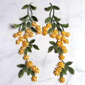 May include: Two symmetrical embroidered floral appliqués featuring vibrant yellow flowers with small white centres and detailed green leaves on stems. These decorative patches are arranged in a trailing vine pattern, showcasing intricate needlework.