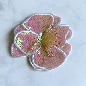 May include: A flower-shaped patch featuring iridescent pink sequins layered on fabric petals with white stitched edges. The center is adorned with a cluster of long, thin, pale yellow and pink glass beads extending outward like stamens.