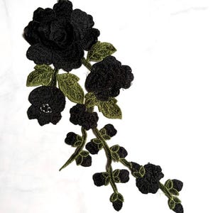 Può includere: Un'applicazione ricamata che presenta due grandi rose nere e un fiore nero più piccolo, collegati da steli e foglie verde oliva. Il design include intricati dettagli in filo sui petali e sul fogliame, creando un motivo floreale.