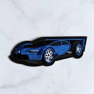 May include: An embroidered patch featuring a sleek blue sports car in profile. The design showcases a vibrant blue body with black detailing, white multi-spoke wheels, and a low-profile aerodynamic shape. The entire car is outlined in black thread.