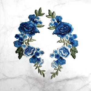 May include: Two symmetrical floral embroidery patches featuring deep blue and light blue roses, smaller blue blossoms, and olive green leaves. The intricate threadwork creates a detailed botanical design suitable for fabric application.