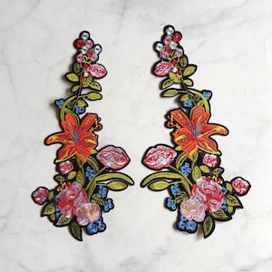 May include: Two symmetrical floral embroidered patches featuring vibrant orange lilies, pink roses, and small blue flowers with green leaves and stems. Each patch has a black border and a detailed stitched design, showcasing a variety of colorful blossoms.