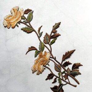 Puede incluir: Aplicación floral bordada con dos rosas de color amarillo dorado con costuras detalladas. Las hojas son una mezcla de verde y marrón, unidas a tallos marrones. Ideal para embellecer ropa o manualidades.