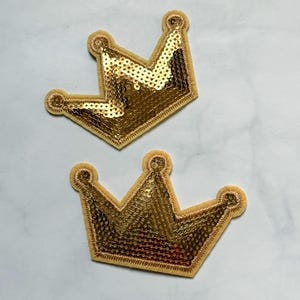 May include: Two gold sequined crown patches. The crowns have a light tan border and are filled with shiny gold sequins. The patches are designed with three points and three rounded edges. The crowns are laying on a white surface.