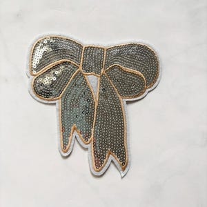 Peut inclure: Un écusson décoratif en forme de nœud avec des paillettes argentées et une bordure dorée. Le nœud a un dos en feutre blanc et est conçu pour l'embellissement de vêtements ou d'accessoires. Les paillettes captent la lumière, créant un effet scintillant.