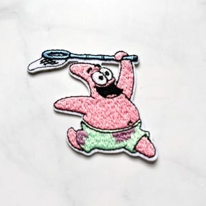 Può includere: Toppa ricamata con Patrick Stella di Spongebob Squarepants. Il personaggio rosa a forma di stella marina tiene una rete blu sopra la testa. Indossa pantaloncini verdi e viola e ha un'espressione felice.