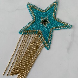 May include: A turquoise star-shaped embellishment with a black star in the center. The star is outlined with gold and clear rhinestones, and has gold beaded fringe hanging from the bottom. The star is on a white surface.