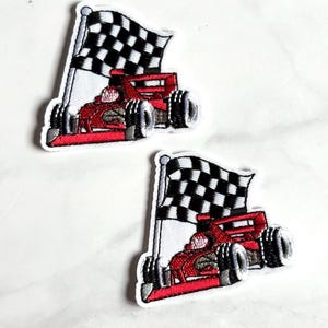 May include: Two embroidered patches featuring a red race car positioned in front of a black and white checkered flag. The patches have a white border and depict a detailed racing vehicle with black tires and a prominent spoiler on the rear.