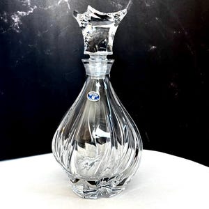 May include: A clear crystal decanter with a decorative stopper. The decanter has a bulbous body with vertical, textured lines. The stopper is a unique, angular shape. A small, blue and white label is affixed to the decanter.