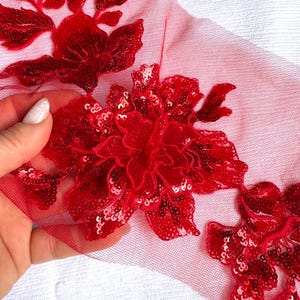 May include: A red floral applique featuring intricate embroidery and shimmering sequins on a sheer red mesh fabric. The design consists of three distinct flower motifs with layered petals, creating a textured and dimensional appearance on the netting.