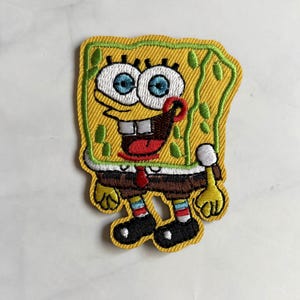 May include: An embroidered patch featuring the cartoon character SpongeBob SquarePants. The character is yellow and rectangular with large white eyes, blue irises, a wide smile, a red tie, brown pants, and black shoes with white accents.