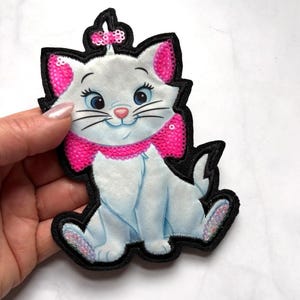 May include: Embroidered patch featuring a white cartoon cat with pink accents on the ears and a pink bow. The cat has blue eyes, a pink nose, and a pink sequined scarf. The patch is outlined in black.