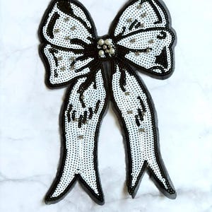May include: A decorative bow appliqué featuring white sequins and black beaded outlines. The centre knot is embellished with a cluster of small white pearls, and additional scattered pearls are placed across the bow loops and long trailing tails.