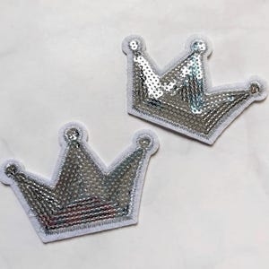 May include: Two silver sequined crown patches with white felt backing. The crowns have a classic five-point design and are outlined with silver stitching. The sequins reflect light, creating a sparkling effect. These patches are ideal for embellishing clothing or accessories.