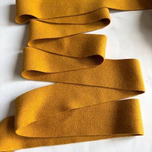 May include: Mustard yellow fabric ribbon, arranged in a flowing pattern. The ribbon has a textured appearance, likely woven. The background is a plain white surface. The ribbon is suitable for crafts.