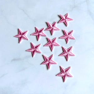 Puede incluir: Un conjunto de diez parches pequeños con forma de estrella de cinco puntas en un tono rosa vibrante. Cada parche presenta un bordado detallado con una superficie texturizada en relieve y un punto central distintivo, dispuestos en triángulo.