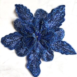 May include: A royal blue floral applique featuring intricate embroidery, shimmering sequins, and delicate beadwork. The flower shape has multiple layered petals with scalloped edges, creating a textured, three-dimensional appearance.