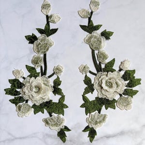 Puede incluir: Dos parches bordados florales simétricos con rosas y capullos en color blanco roto, acompañados de hojas y tallos en verde oliva oscuro. El intrincado bordado muestra pétalos superpuestos y follaje detallado en estos apliques.