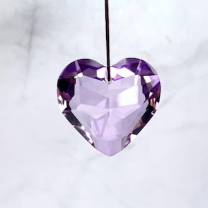 May include: A faceted, heart-shaped pendant made of translucent light purple crystal. The pendant features a hole at the top center and is suspended by a thin, dark brown cord, showcasing geometric cuts that reflect light across its surface.