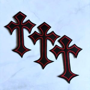 May include: Three embroidered patches in the shape of a cross. Each patch features a black center with a vibrant red stitched border outlining the cross shape. The patches have a textured fabric appearance with sharp, pointed edges on all sides.